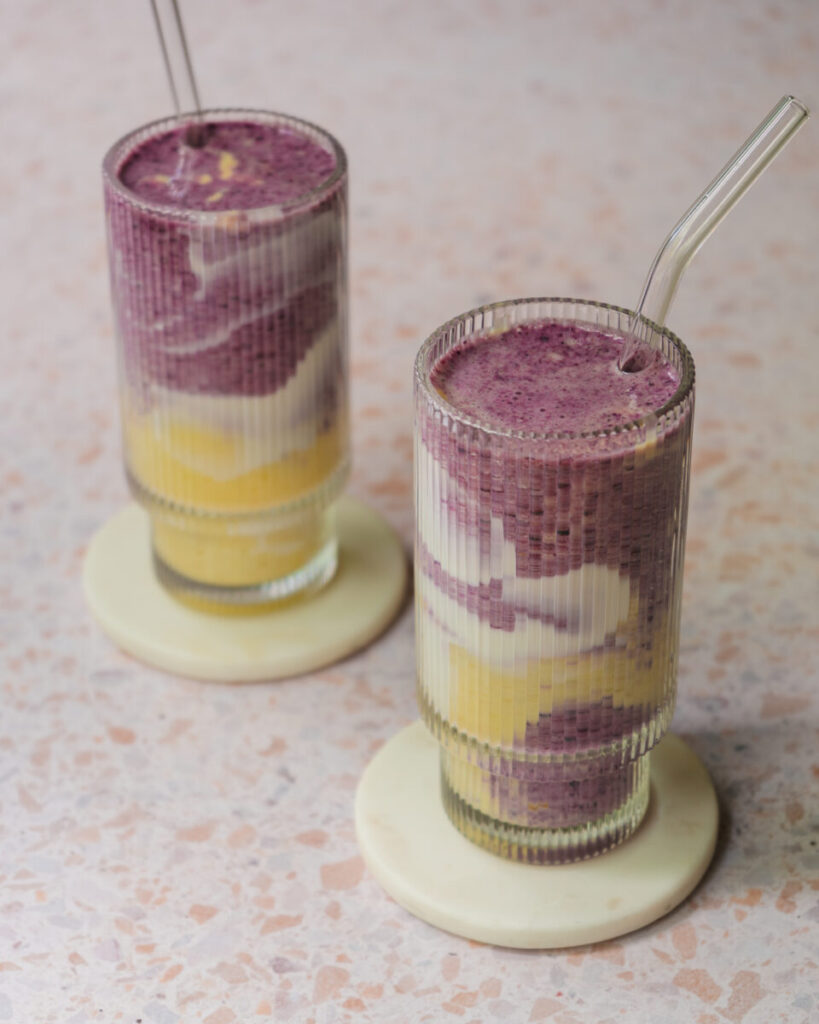 Smoothie marbré aux bleuets et à la mangue servi dans un verre, avec des tourbillons violets et orangés et une décoration de yogourt sur les parois.